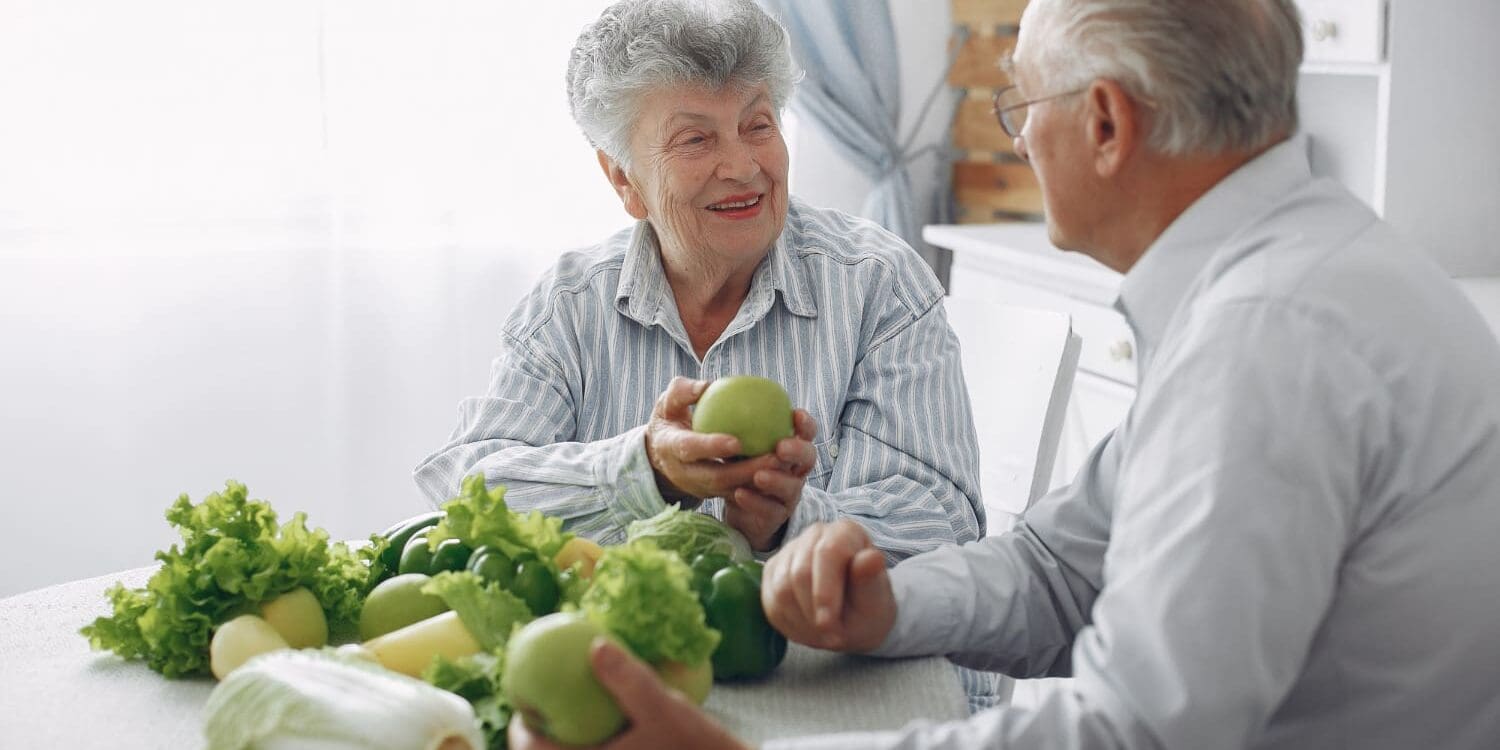 Twee mensen met artrose zitten aan een tafel met groenten die veel vitamine k bevatten.